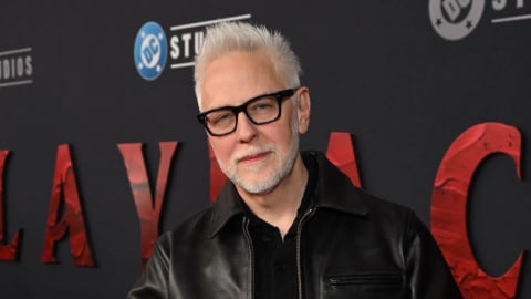 The image shows filmmaker James Gunn attending the premiere of "Clayface." Gunn is captured from the chest up, positioned slightly off-center to the right, against a dark gray backdrop featuring the text "CLAYFACE" in large, textured red capital letters, alongside the DC Studios logo. He is smiling faintly, looking directly at the camera, and has short, styled white hair with a light gray beard and mustache. He is wearing thick-rimmed black eyeglasses, a black collared button-down shirt, and a classic black leather jacket with a pointed collar. The lighting highlights the texture of his jacket and hair, creating a sharp contrast against the dark background.
