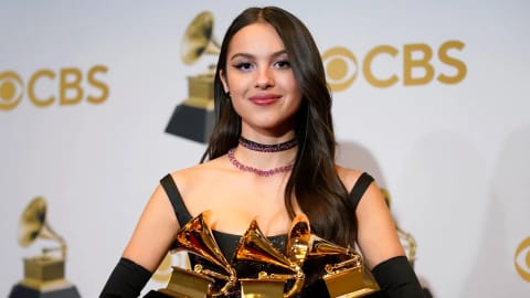 This image shows American singer-songwriter Olivia Rodrigo posing with three Grammy awards at the 64th Annual Grammy Awards press room on April 3, 2022, in Las Vegas, Nevada. She is wearing a custom black corseted gown by Vivienne Westwood, paired with matching opera gloves, a pink-and-black choker necklace, and a sharp winged eyeliner look.