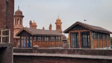 A high-angle, outdoor shot captures the historic red brick rooftop of the National College of Arts (NCA) in Lahore, featuring two distinct, vintage wooden cabins alongside towering Mughal-style architectural elements beneath a hazy, overcast sky. The foreground and middle ground are dominated by weathered, dark red brick structures, including a thick boundary wall that separates the immediate rooftop area from the background. Built upon this rooftop are two elongated, rustic wooden pavilions or cabins, each characterized by dark timber paneling, multiple small-paned glass windows reflecting a hint of blue from an interior object, and pitched roofs covered in classic terracotta tiles with ornate, scalloped wooden trim along the eaves. Rising majestically behind these wooden structures are several iconic, reddish-brown terracotta domes and minarets belonging to a historic architectural complex, their elegant curves and intricate spires standing out against the pale, muted grey and white sky where a flock of small birds can be seen scattered in flight. The left side of the frame features a stark, vertical red brick wall section with exposed piping, adding a raw, structural texture to the composition, while the soft, diffused daylight casts gentle shadows across the aged masonry and wooden surfaces, highlighting the rich cultural heritage and academic atmosphere of the fine arts department's outdoor space.