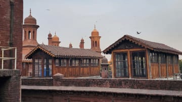 A high-angle, outdoor shot captures the historic red brick rooftop of the National College of Arts (NCA) in Lahore, featuring two distinct, vintage wooden cabins alongside towering Mughal-style architectural elements beneath a hazy, overcast sky. The foreground and middle ground are dominated by weathered, dark red brick structures, including a thick boundary wall that separates the immediate rooftop area from the background. Built upon this rooftop are two elongated, rustic wooden pavilions or cabins, each characterized by dark timber paneling, multiple small-paned glass windows reflecting a hint of blue from an interior object, and pitched roofs covered in classic terracotta tiles with ornate, scalloped wooden trim along the eaves. Rising majestically behind these wooden structures are several iconic, reddish-brown terracotta domes and minarets belonging to a historic architectural complex, their elegant curves and intricate spires standing out against the pale, muted grey and white sky where a flock of small birds can be seen scattered in flight. The left side of the frame features a stark, vertical red brick wall section with exposed piping, adding a raw, structural texture to the composition, while the soft, diffused daylight casts gentle shadows across the aged masonry and wooden surfaces, highlighting the rich cultural heritage and academic atmosphere of the fine arts department's outdoor space.