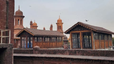 A high-angle, outdoor shot captures the historic red brick rooftop of the National College of Arts (NCA) in Lahore, featuring two distinct, vintage wooden cabins alongside towering Mughal-style architectural elements beneath a hazy, overcast sky. The foreground and middle ground are dominated by weathered, dark red brick structures, including a thick boundary wall that separates the immediate rooftop area from the background. Built upon this rooftop are two elongated, rustic wooden pavilions or cabins, each characterized by dark timber paneling, multiple small-paned glass windows reflecting a hint of blue from an interior object, and pitched roofs covered in classic terracotta tiles with ornate, scalloped wooden trim along the eaves. Rising majestically behind these wooden structures are several iconic, reddish-brown terracotta domes and minarets belonging to a historic architectural complex, their elegant curves and intricate spires standing out against the pale, muted grey and white sky where a flock of small birds can be seen scattered in flight. The left side of the frame features a stark, vertical red brick wall section with exposed piping, adding a raw, structural texture to the composition, while the soft, diffused daylight casts gentle shadows across the aged masonry and wooden surfaces, highlighting the rich cultural heritage and academic atmosphere of the fine arts department's outdoor space.