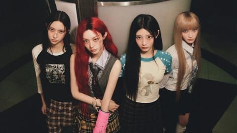 A wide, overhead shot captures the four members of the K-pop group ILLIT standing closely together, leaning against a curved architectural structure adorned with an illustration. They all look directly upward into the camera with serious, intense expressions. The member on the far left wears a black graphic tee with a "X-girl" print paired with dark plaid pants. The second member has vibrant red hair and a pink cast on her left arm, dressed in a grey sweater vest over a white collared shirt and a yellow plaid pleated skirt. The third member sports a cream-colored raglan tee with blue star-patterned sleeves and a dark plaid skirt. The member on the far right has long blonde hair with blunt bangs, wearing a striped button-up shirt, a tie, and a dark skirt. The overall aesthetic is moody and nostalgic, cast in warm, muted lighting.