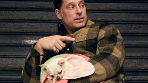 This image captures actor Joe Manganiello alongside his famous rescue Chihuahua-Pomeranian mix, Bubbles, during a Netflix promotional interview. Actor Joe Manganiello sits in front of a dark, horizontally corrugated industrial metal shutter, wearing an olive green and black plaid flannel button-down shirt while cradling a tiny white Chihuahua swaddled in a pink and green towel. His hair is slicked back into a neat bun, and his gaze is directed to the right with a slightly open-mouthed, animated expression. He points his right index finger across his chest toward his left shoulder, showcasing a large, ornate gold skull ring on his ring finger and a thin gold chain bracelet on his wrist. The small, light-furred Chihuahua rests quietly in his left arm, its head tilted downward with a gentle, calm demeanor amid the colorful pastel fabrics.