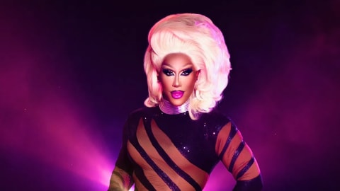 The image features drag artist Kori King striking a powerful pose in a stunning Halloween transformation that channels iconic superstar RuPaul. She wears a form-fitting, sparkling black bodysuit detailed with dramatic, sheer mesh diagonal cutouts that wrap around her torso and legs, creating a striking optical illusion. Her ensemble is elevated by a voluminous, meticulously styled platinum blonde wig, impeccably contoured makeup featuring a bold purple lip, and a metallic silver statement choker. Bathed in a dramatic, hazy magenta spotlight against a dark background, the entire look serves as a flawless, high-glamour tribute to the Queen of Drag.