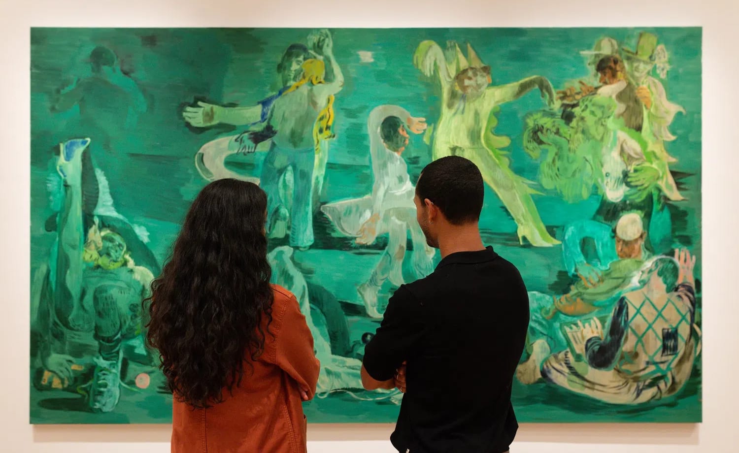 A wide gallery shot captured inside The Courtauld Gallery shows two gallery visitors—a woman with long, dark curly hair wearing an orange jacket on the left and a man in a black long-sleeved shirt on the right—standing with their backs to the camera as they closely study a massive figurative painting. The artwork, titled Ghost Ball (2023) by celebrated artist Salman Toor, dominates the frame with its signature, surreal palette of deep emerald and absinthe greens. Across the vast canvas, a series of loose, fluid, and elongated figures are depicted in various expressive, theatrical poses that suggest a dreamlike nocturnal gathering or party. The figures—some wearing whimsical caps or costumed elements, others lounging or dancing—inhabit a stylized, safe nocturnal environment that evokes themes of community, intimacy, and queer South Asian experiences. The bright gallery lighting illuminates the clean white wall surrounding the massive canvas, casting a subtle warmth on the shoulders of the spectators engrossed in the scene.