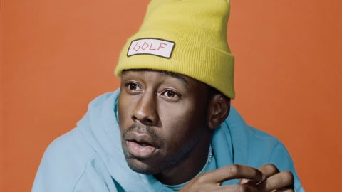 A close-up portrait of the artist Tyler, The Creator against a solid, vibrant orange backdrop. He is wearing a bright yellow knit beanie adorned with a white rectangular patch featuring the word "GOLF" in pink, stylized lettering. He sports a light blue hoodie with embroidered text partially visible near the collar. His expression is contemplative with an open-mouthed, slightly surprised look as he gazes off-camera to his left, his hands raised in front of him with a silver ring visible on his pinky finger.