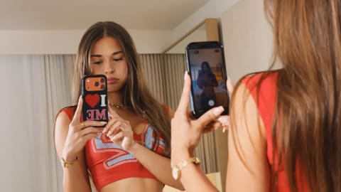 A young woman with long, straight brown hair, identified as Sophie Rain, stands in a brightly lit bedroom facing a large mirror while capturing a mirror selfie with her smartphone. She is wearing a sleeveless red and grey argyle patterned crop top that exposes her midriff, paired with blue and white gingham patterned pajama shorts over a red bikini bottom or underwear. Her phone features a prominent black case with a graphic design reading "I ❤️ ME". Her reflection in the mirror shows her focused expression as she frames the shot, while the foreground captures her from behind, highlighting the texture of her hair and the casual, relaxed style of her outfit.