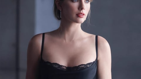 A close-up promotional image of singer-songwriter Taylor Swift gazing thoughtfully to the right. She wears her hair gathered in a soft updo with structured, side-swept bangs framing her face. Her makeup is minimalist with a subtle brown smoky eye and soft coral-pink lipstick. She is wearing a slinky, low-cut black satin camisole slip dress featuring a delicate black lace trim along the curved neckline. Her shoulders are bare except for thin spaghetti straps, and dainty silver drop earrings catch the light. The background consists of modern, muted gray concrete pillars with soft, diffused lighting filtering in from the side.