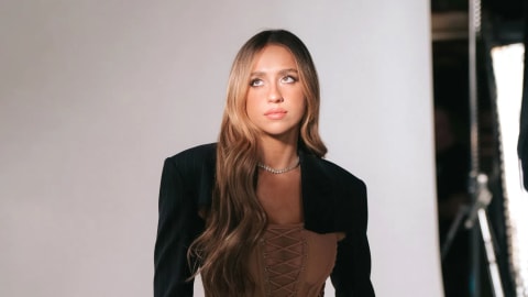 Canadian singer, songwriter, and dancer Tate McRae poses during a studio photoshoot, looking off-camera with a contemplative expression. Her long, wavy caramel-brown hair cascades over her shoulder, complementing her distinct early-2000s inspired outfit. She wears a structured, cropped black blazer shrugged over a tan lace-up corset top, paired with low-rise, oversized beige cargo pants that reveal a glimpse of patterned underwear at the waistband. A simple diamond tennis necklace completes the look against a clean, lit studio backdrop featuring photographic equipment in the soft-focus background.