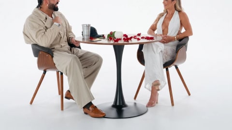 The image captures a professionally lit studio setting featuring Canadian rapper and global superstar Drake sitting across a round, dark-wood bistro table from a woman during a speed dating sequence. The table is distinctively decorated with scattered red rose petals. Drake is positioned on the left side of the frame, styled in matching loose-fitting, beige or tan utility streetwear pants and a matching jacket over a white undershirt. His hair is neatly braided into cornrows, and he is wearing brown leather loafers with white socks, leaning slightly forward with a broad smile while gesturing with his right hand toward his companion.Seated opposite him on the right is a woman with blonde hair, wearing a deeply plunging, halter-neck white dress featuring a sheer lace overlay skirt. Both individuals are sitting in modern mid-century styled armchair seats equipped with dark grey padded cushions and curved walnut wood frames supported by tapered dowel legs. The backdrop is entirely flat and seamless white studio space, creating a stark, high-contrast visual environment that draws all attention to the interactive date setup at the center.