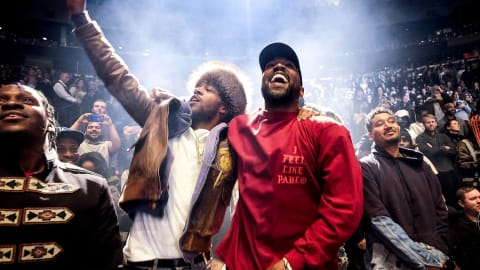 An image captures Kanye West and Kid Cudi sharing a moment of triumph during the listening party for West’s album The Life of Pablo at Madison Square Garden. Standing side-by-side in the midst of a packed, brightly illuminated stadium crowd, West laughs broadly while wearing a red long-sleeved shirt with the text "I FEEL LIKE PABLO" printed on the chest and a black cap. Beside him, Kid Cudi extends an arm high into the air while singing or shouting enthusiastically, dressed in a white t-shirt under a heavily layered brown jacket with a prominent fur-trimmed hood. To their left, rapper Pusha T is visible in profile looking upward, and behind them, an expansive audience fills the arena seats, captured under dramatic spotlighting that creates a hazy, energetic atmosphere.