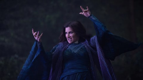 An official production still from Marvel Television's "Agatha All Along" showcases actress Kathryn Hahn reprising her role as the formidable witch Agatha Harkness. Captured in a medium shot against a dimly lit, shadowy forest backdrop, she embodies dark, mystical power while dressed in her signature elaborate witch robes—a complex ensemble featuring rich shades of deep blue and purple with heavily textured, pleated, and billowing fabric that gathers at her waist. Her dark hair flows loosely as she strikes a theatrical, spellcasting pose with both arms raised, her claw-like hands held high with fingers dramatically bent, exposing dark nail polish. She displays an intense, fierce facial expression with bared teeth and a calculating gaze, evoking a sinister and chaotic energy characteristic of her supernatural persona. The cool, moody lighting filters subtly through the background, casting deep shadows over the scenery and accentuating the intricate contours of her costume to elevate the overall eerie, witchy aesthetic of the scene.