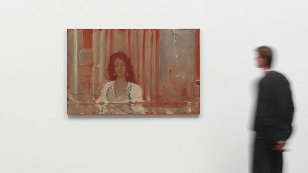An installation photograph from White Cube Paris showcases a rectangular, horizontally oriented figurative painting by Pakistani artist Suleman Aqeel Khilji. The artwork is mounted centrally on a large, pristine white gallery wall. Executed with a muted, warm palette dominated by earth-toned reds, terracottas, and hazy greys, the dreamlike composition depicts a woman with voluminous dark curly hair sitting behind what appears to be a counter or reflective surface. She wears a white, low-cut blouse or dress with dark trim along the neckline. The background features hazy vertical bands resembling curtains or architectural pillars, rendered in semi-translucent layers of pigment that evoke a sense of a faded memory or an excavated artifact. To the right of the canvas, a person wearing a black long-sleeved shirt and dark trousers stands on a polished concrete gallery floor, captured in motion blur as they look toward the left.