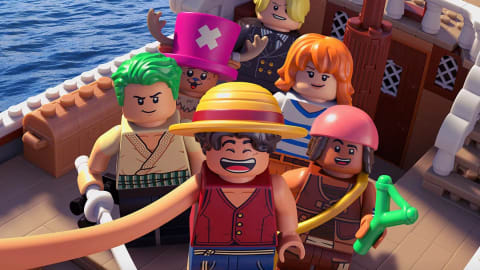 An official promotional poster styled as a Lego adaptation of the live-action Netflix series, featuring several iconic Straw Hat Pirates characters assembled on the deck of their ship, the Going Merry. A promotional poster styled as a LEGO video game or animated special for the Netflix live-action series adaptation of One Piece. The image is set on the deck of a detailed LEGO version of the pirate ship, the Going Merry, sailing across a vibrant blue plastic brick ocean. In the foreground, six iconic Straw Hat Pirates are depicted as LEGO minifigures. At the center is Monkey D. Luffy, wearing his signature straw hat and red vest, smiling widely as he stretches his long LEGO arm forward to take a selfie. Flanking him are Roronoa Zoro with green hair and two swords, and Usopp holding a green slingshot. Behind them stand Tony Tony Chopper wearing his pink top hat, Sanji with blonde hair over one eye in a black suit, and Nami in a blue and white striped shirt. In the background, a giant grey brick-built whale, Laboon, breaches out of the water. Text at the top reads "BUILT DIFFERENTLY," and the bottom features the "LEGO" logo alongside the "ONE PIECE" skull-and-crossbones logo, captioned with "A NETFLIX LIMITED SERIES" and "ONLY ON NETFLIX | NOW PLAYING."