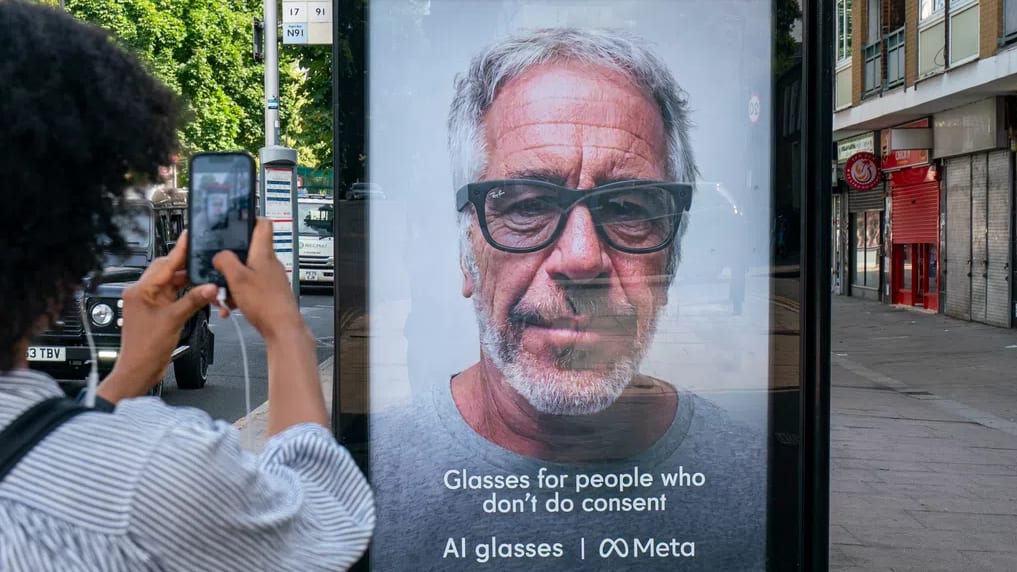 The image captures a street scene focused on a controversial mock advertisement displayed on a digital bus stop shelter or billboard. In the foreground on the left, a person with dark, curly afro hair, wearing a striped shirt, holds up a smartphone to take a picture of the ad. The advertisement features a large, close-up, grayscale portrait of convicted sex offender Jeffrey Epstein wearing a pair of thick, dark-rimmed smart glasses. Text printed in white at the bottom of the ad reads, "Glasses for people who don't do consent" followed by "AI glasses | ♾️ Meta", parodying Meta's Ray-Ban smart glasses marketing campaign. The ad is reflected in its glass casing, showing hints of the outdoor environment. The background reveals an urban sidewalk, a closed storefront with a red security shutter, and vehicles parked on the street under daylight.