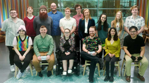 A group portrait of the cast and crew from the Australian documentary series, The Assembly. The group consists of fifteen individuals arranged in two rows inside a modern room with a backdrop of colorful, vertical stripes. In the back row, eight people stand side by side: a man in a light green striped shirt, a woman in a red V-neck sweater, a man with a large red beard wearing a grey sweater, a tall man with glasses in a blue button-down shirt, a man in a white polo shirt, a man in a patterned pink shirt, host Leigh Sales in a dark blue velvet blazer over a white shirt, a woman with long dark hair, a woman with long blonde hair, and a man in a white short-sleeve shirt with a leaf pattern. In the front row, six individuals sit facing forward: a person with colorful rainbow argyle vest and pink beanie, a man in a green shirt and khaki pants holding a sign, a woman with glasses and a sparkling jacket sitting in a motorized wheelchair, a man in a tropical floral shirt, a woman in a yellow wrap top, and a man in a black polo shirt and beige pants wearing arm compression sleeves.