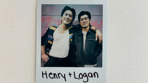 The image features a Polaroid-style photo stuck to a plain white wall, showcasing two smiling young men standing shoulder to shoulder. The person on the left wears a white tank top under an open, colorful patterned cardigan, while the person on the right wears a dark jacket and glasses, with the name "Henry + Logan" handwritten in black marker across the bottom white border.