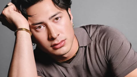 The image captures a striking studio portrait of Hudson Williams, looking directly into the camera with a calm, introspective expression while leaning forward against a dark surface. His dark, wavy hair is parted down the middle and styled elegantly, framing a sharp face highlighted by warm lighting against a neutral, slate-gray backdrop. He is wearing a short-sleeved, taupe-brown knit polo shirt with a fine textured ribbed collar, showing a partial tattoo on his right inner forearm. His right hand rests against his temple, showcasing an ornate gold bracelet and small gold hoop earrings, while his left arm rests horizontally across the bottom of the frame, drawing attention to a luxury Bvlgari watch with a gold case and a black leather strap.