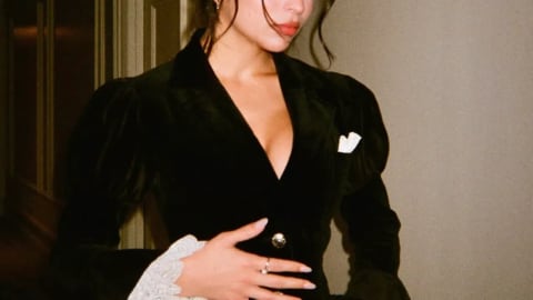 A flash-photographed, medium shot of American actress Josie Totah looking slightly to her right while posing indoors against a neutral, beige wall. She has dark hair styled in an elegant updo with loose, wavy face-framing tendrils. She wears a structured black velvet blazer or mini dress featuring a deep V-neckline, prominent puffed shoulders, and ornate gold buttons fastening the front. A small white pocket square accents the left side of the chest. The long sleeves flare dramatically at the wrists into tiered, voluminous white lace ruffles. She rests her manicured hands with long, light-colored nails and multiple metallic rings over her torso, over thin black sheer tights. A ventilation grate is visible on the upper right wall in the softly lit background.