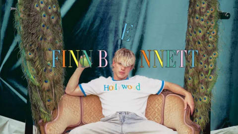 The actor Finn Bennett poses for the cover of CYNOSURE magazine, sitting on an ornate gold velvet sofa flanked by taxidermy peacocks against a dark teal backdrop. He is wearing a white T-shirt with blue ringer trim that reads "Hollywood," cuffed light-wash denim jeans, and tan leather cowboy boots, lounging with his legs spread wide and one arm resting along the back of the couch. The magazine's title "CYNOSURE" is stylized in a colorful, metallic serif font at the top, while his name "FINN BENNETT" is boldly overlaid across the middle of the composition in gold and green text.