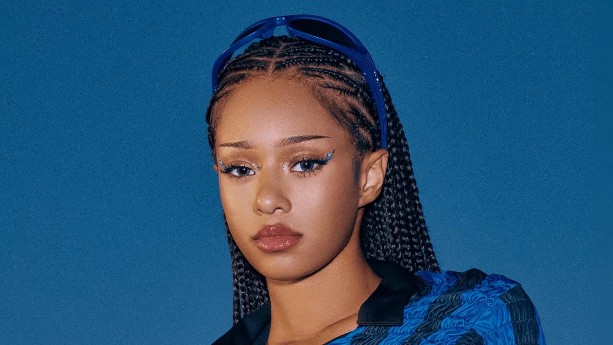 The image shows a portrait of model and content creator Manon Bannerman set against a solid blue background, styled in a sporty, Y2K-inspired aesthetic. She wears an Inter Milan football shirt featuring vertical blue and black stripes, a black collar, and the white "Pirelli" sponsor logo across the chest, uniquely updated with a crinkled, textured popcorn fabric. Her hair is styled in neat, symmetrical cornrow braids extending to the back, with blue-framed sunglasses resting on top of her head. Her makeup is sharp, highlighting soft brown lipstick, winged graphic eyeliner adorned with small silver metallic accents near the eyes, and well-defined brows. A portrait showcases Manon Bannerman wearing a vintage-style Inter Milan soccer jersey modified into a smocked, texture-rich long-sleeve top featuring the classic white and yellow Pirelli front sponsor logo against vertical blue and black stripes. She is styled with neat, long box braids drawn straight back, dark sunglasses propped up on her head as a hairband, subtle cat-eye eyeliner, and neutral lip gloss, all set against a solid deep blue background.
