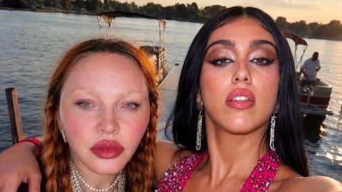 The image shows Madonna and her daughter, Lourdes Leon, standing closely together on a dock beside a body of water at sunset. Madonna, on the left, features long, reddish-blonde braided hair, bleached eyebrows, prominent pink blush, and bold red lipstick. She is wearing a black mesh top, a wide buckled belt, and layered silver chain necklaces, including a large cross pendant. Lourdes Leon, on the right, has long, straight black hair, dramatic eye makeup, and glossy lips. She wears a vibrant, embellished pink halter top with matching wrap detailing, raising her arm to take the selfie and naturally revealing her unshaved underarm. In the background, calm water stretches out with a pontoon boat docked nearby and trees lining the distant shore under a soft evening sky.