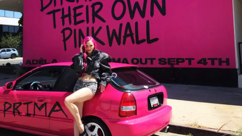 The image shows a vibrant promotional scene featuring artist ADÉLA posing on a custom hot pink car in front of the iconic Paul Smith Pink Wall on Melrose Avenue in Los Angeles. The wall features a bold, graffiti-style black inscription that reads, "THE BADDEST BITCHES HAVE THEIR OWN PINK WALL," alongside text underneath announcing her project "PRIMA OUT SEPT 4TH." ADÉLA matches the aesthetic with hot pink streaks in her hair, denim shorts, and a black puffer jacket, while sitting on a pink hatchback that has "PRIMA" spray-painted across the side door.