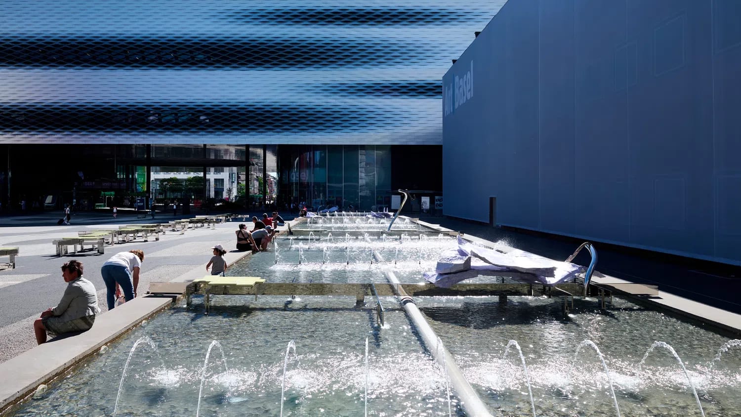 The image features a large-scale sculptural installation by artist Nairy Baghramian titled Modèle vivant (S’empilant) (2026), which is currently situated in the Messeplatz fountain in front of the Messe Basel exhibition hall in Switzerland.