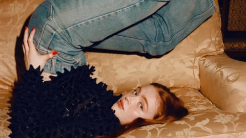 A young woman with long, vibrant red hair poses playfully upside down on a plush, gold damask-patterned sofa, her legs elevated high in the air while wearing mid-wash blue denim jeans paired with white, patterned crew socks. She wears an intricate, heavily textured navy or black long-sleeved top covered in small, 3D fabric embellishments or petal-like structures that give it a unique tactile depth. Resting on her back with her head tilted toward the camera, she gazes directly forward with a soft, neutral expression, accented by a bright red manicure on her hand resting near her hip. The indoor setting features a vintage aesthetic with warm, subdued lighting, highlighted by a glowing wall sconce in the background and a classic wooden paneled door, casting dramatic shadows that enhance the cozy, editorial feel of the photograph.