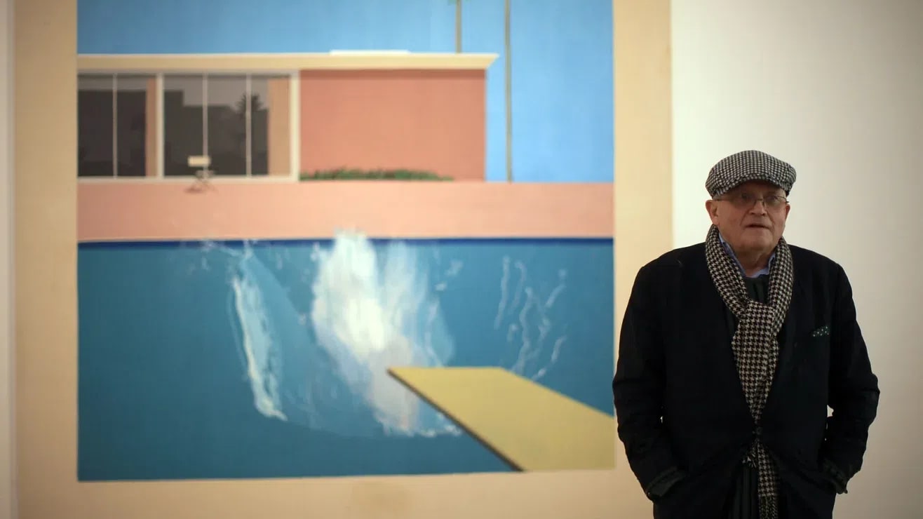 A wide, eye-level shot captures British artist David Hockney standing to the right of his famous large-scale pop art painting, A Bigger Splash (1967), which is mounted on a plain white gallery wall. The painting itself takes up the majority of the left and center portions of the frame. It depicts a modernist, flat-roofed house with floor-to-ceiling glass windows under a bright, clear blue sky, flanked by two tall, thin palm trees. In the foreground, a yellow diving board extends from the bottom right into a swimming pool of turquoise water, where a massive, chaotic splash of white and light blue water erupts, indicating someone has just dived in. The entire painting is bordered by a wide, unpainted canvas margin. To the right, David Hockney stands in a three-quarter view, looking slightly past the camera to the left. He is an elderly man with glasses, wearing a dark navy blazer, a black and white houndstooth scarf draped around his neck, and a matching houndstooth flat cap. His hands are tucked into his pockets, and his expression is neutral and contemplative. The lighting is soft and even, typical of a gallery setting, casting subtle shadows behind him.
