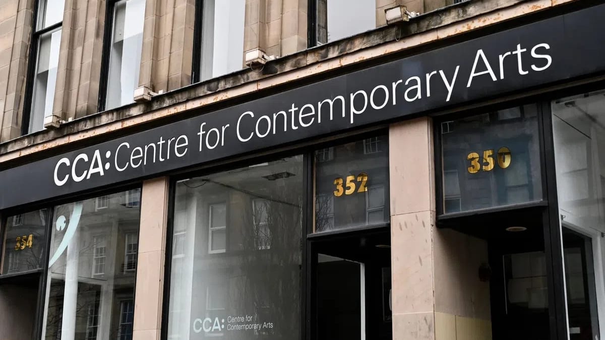 A black sign featuring the words "CCA: Centre for Contemporary Arts" in bold white lettering hangs prominently above the entrance of a stone building with large, dark-framed display windows and glass doors reflecting surrounding architectural details. A horizontal, eye-level shot captures the exterior entrance of the Centre for Contemporary Arts (CCA) building, focusing on a long, sleek black signboard with the institution's name written in crisp, clean white sans-serif font. Below the sign, large transparent glass windows and glass entrance doors are set within a pale stone facade, reflecting the outlines of historical buildings across the street, while gold numbers "352" and "350" are visible on the glass panels above the doorways.