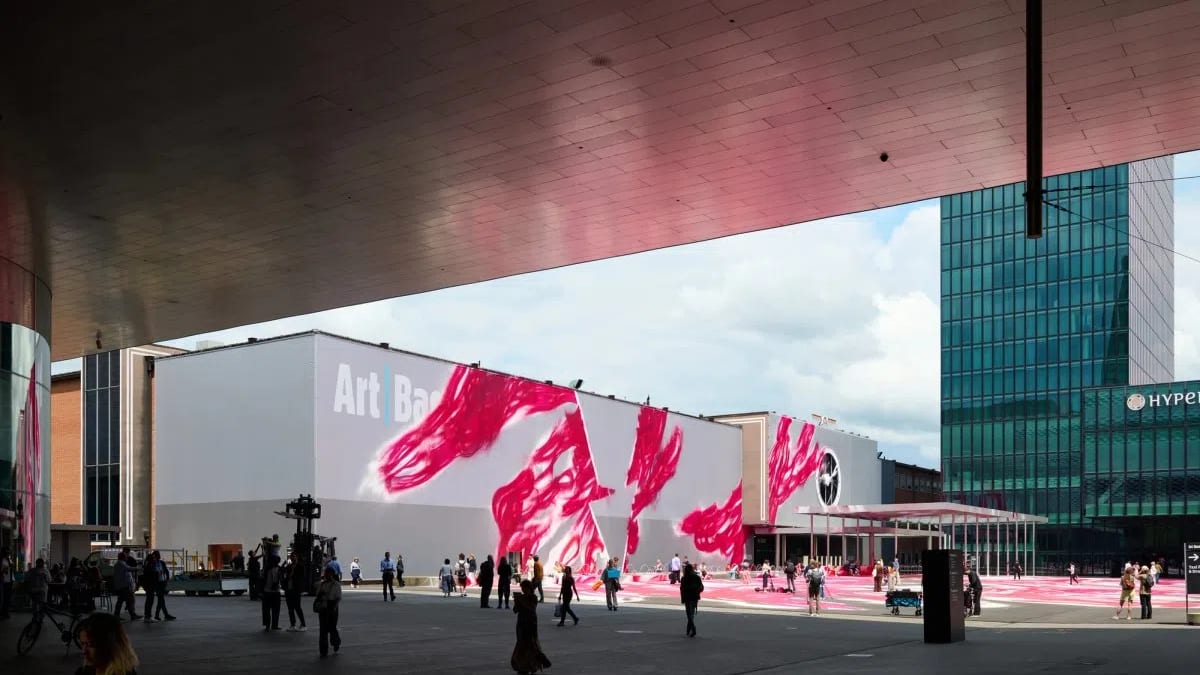A wide outdoor view of Messeplatz during the Art Basel fair, captured from beneath the shelter of a large, metallic architectural overhang. In the center, a large white exhibition hall prominently features the words "Art Basel" alongside an abstract installation consisting of giant, vibrant pink paintbrush strokes that trail down the facade and spread across the open plaza floor. People wander through the open square, while a modern green glass skyscraper rises into the cloudy sky on the right side of the frame.