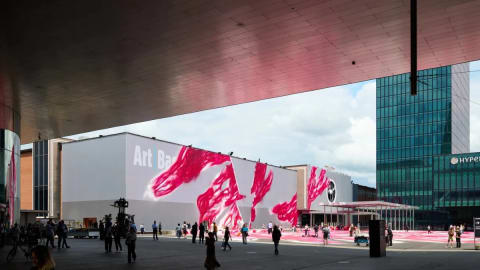 A wide outdoor view of Messeplatz during the Art Basel fair, captured from beneath the shelter of a large, metallic architectural overhang. In the center, a large white exhibition hall prominently features the words "Art Basel" alongside an abstract installation consisting of giant, vibrant pink paintbrush strokes that trail down the facade and spread across the open plaza floor. People wander through the open square, while a modern green glass skyscraper rises into the cloudy sky on the right side of the frame.