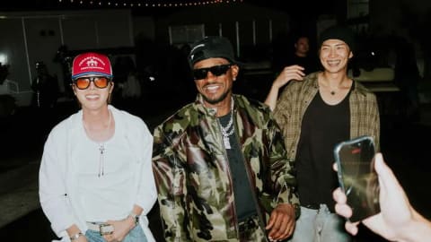 An image captured at night shows BTS members J-Hope and RM smiling and laughing alongside American singer Chris Brown. On the left, J-Hope wears a vibrant red trucker hat, tinted sunglasses, a loose white button-up shirt, and baggy light-wash jeans while flashing a wide smile. In the center, Chris Brown sports a camo-print leather jacket over a black top, a black baseball cap, dark sunglasses, and chunky silver chains. To the right, RM beams in a relaxed, casual outfit consisting of an open brown plaid flannel shirt over a black t-shirt and a dark beanie. The background shows an outdoor venue setting illuminated by string lights, with a hand holding up a smartphone in the foreground to snap a photo of the trio. 