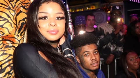 A medium close-up shot captures media personalities Chrisean Rock and Blueface inside a crowded, dimly lit nightclub venue. Chrisean Rock stands prominently on the left side of the frame, looking directly into the camera with long, dark straight hair draped over her shoulder and a subtle smile. She wears a long-sleeved black top and a silver metallic chain bracelet on her wrist. Behind her left shoulder, Blueface stands slightly lowered in the frame, wearing a blue jacket with his characteristic facial tattoos visible. The background is filled with a dense crowd of clubgoers behind a metal security barrier, multiple camera flashes illuminating the dark space, neon purple lighting patterns on the ceiling, and a tiger-print patterned garment visible on the far left.