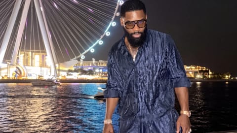 The image features Igho "Tiny" Ubiribo posing confidently on a boat at night, set against the vibrant backdrop of a massive, illuminated Ferris wheel—the Ain Dubai—stretching into the dark sky. He is dressed in a stylish, matching dark navy blue plissé or crinkled two-piece shirt and pants set that subtly catches the surrounding city lights. Accessorizing with large, tinted sunglasses, a thick beard, a classic luxury watch, and a bracelet, he leans casually against the boat's metallic railing. The dark, shimmering water in the foreground reflects the brilliant white and gold neon glows from the observation wheel and the distant coastal skyline, creating a sophisticated and upscale nocturnal atmosphere.