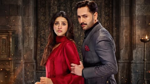 An intense promotional poster features Pakistani actors Danish Taimoor and Anmol Baloch standing close together against a dark, dramatic stone backdrop. On the left, Baloch is captured from the waist up, dressed in a vibrant red traditional outfit with her long dark hair falling over her shoulders, her expression one of worry or concern. On the right, Taimoor stands in profile, looking directly at the camera with a stern, focused gaze. He is wearing a sharp, dark grey tailored suit jacket over a black shirt, with a red pocket square matching her attire. His hand is placed firmly on her arm, suggesting a protective or controlling stance. The atmosphere is elevated by warm lighting, flying sparks, and a classical stone archway with an ornate screen behind them. Drama seriel Nashtar, image by PakistaniArt.