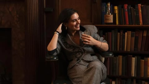 A smiling woman with dark hair sits in a vintage leather armchair in a cozy, dimly lit library, holding a short glass with a beverage. She is wearing a stylish black and white houndstoop skirt suit or pantsuit featuring a dark velvet collar and intricate black swirl detailing on the front. She rests her right hand against her head while looking downward and laughing. In the background, floor-to-ceiling wooden bookshelves are tightly packed with multi-colored antique books, and a bottle of liquor sits on an upper shelf, enhancing the classic, academic atmosphere.