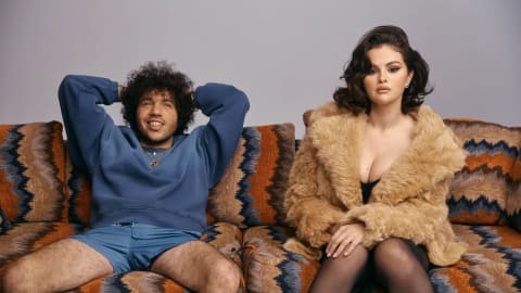 A medium shot of music producer Benny Blanco and singer Selena Gomez sitting side-by-side on a patterned fabric sofa. On the left, Benny Blanco leans back casually with his hands resting behind his head, sporting a full head of curly dark hair, a subtle smile, a blue long-sleeved crewneck sweatshirt, and matching blue shorts. On the right, Selena Gomez sits upright facing forward with a poised, serious expression, her dark hair styled in voluminous waves. She is dressed in a plush, oversized tan faux fur coat draped over a black top, paired with black tights. The sofa features a bold, textured geometric pattern in earthy tones of orange, brown, and black against a plain, neutral light gray studio background.
