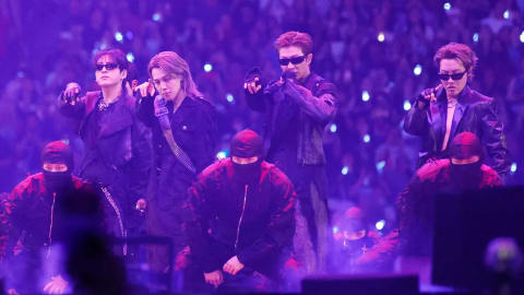 A wide-angle, concert-stage photograph captures four members of the South Korean boy band BTS performing under intense, vibrant purple lighting during a live stadium show. The performers stand side-by-side in a dynamic lineup, wearing stylized, dark edgy clothing, with three of them donning black sunglasses. From left to right, Jungkook gestures forward with his hand, Jimin looks intently toward the audience, RM holds a microphone up to speak while pointing forward, and J-Hope strikes a stylized pose on the right. In front of them, three backup dancers dressed in matching black balaclavas and dark jackets crouch low toward the stage floor. The background is filled with a vast, blurry crowd of fans holding glowing lightsticks, creating an immersive, high-energy concert atmosphere.