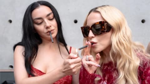 This image captures a multi-generational pop crossover moment featuring English singer-songwriter Charli XCX and American music icon Madonna, mirroring a long-standing creative kinship between the two artists. In the top half, a candid, dimly lit club photograph documents an intimate party gathering from a previous year, showing Charli XCX on the left clad in a sleek black dress holding an unlit cigarette, leaning in toward a blonde Madonna who wears a light pink lace corset top and matching sheer gloves, while a male companion looks on from the right. The bottom half presents a highly publicized modern recreation of that very interaction against a stark, minimalist concrete backdrop. Here, a brunette Charli XCX in a vibrant red lace top holds up a lighter to ignite a cigarette for Madonna, who sports wavy blonde locks, oversized dark tortoiseshell sunglasses, a textured maroon top, and an array of statement rings. The side-by-side comparison highlights not only their evolving personal styles and the shift from raw nightlife documentary to polished promotional aesthetic, but it also underscores the enduring mentor-like bond and mutual artistic admiration shared between the veteran Queen of Pop and the hyperpop trailblazer.