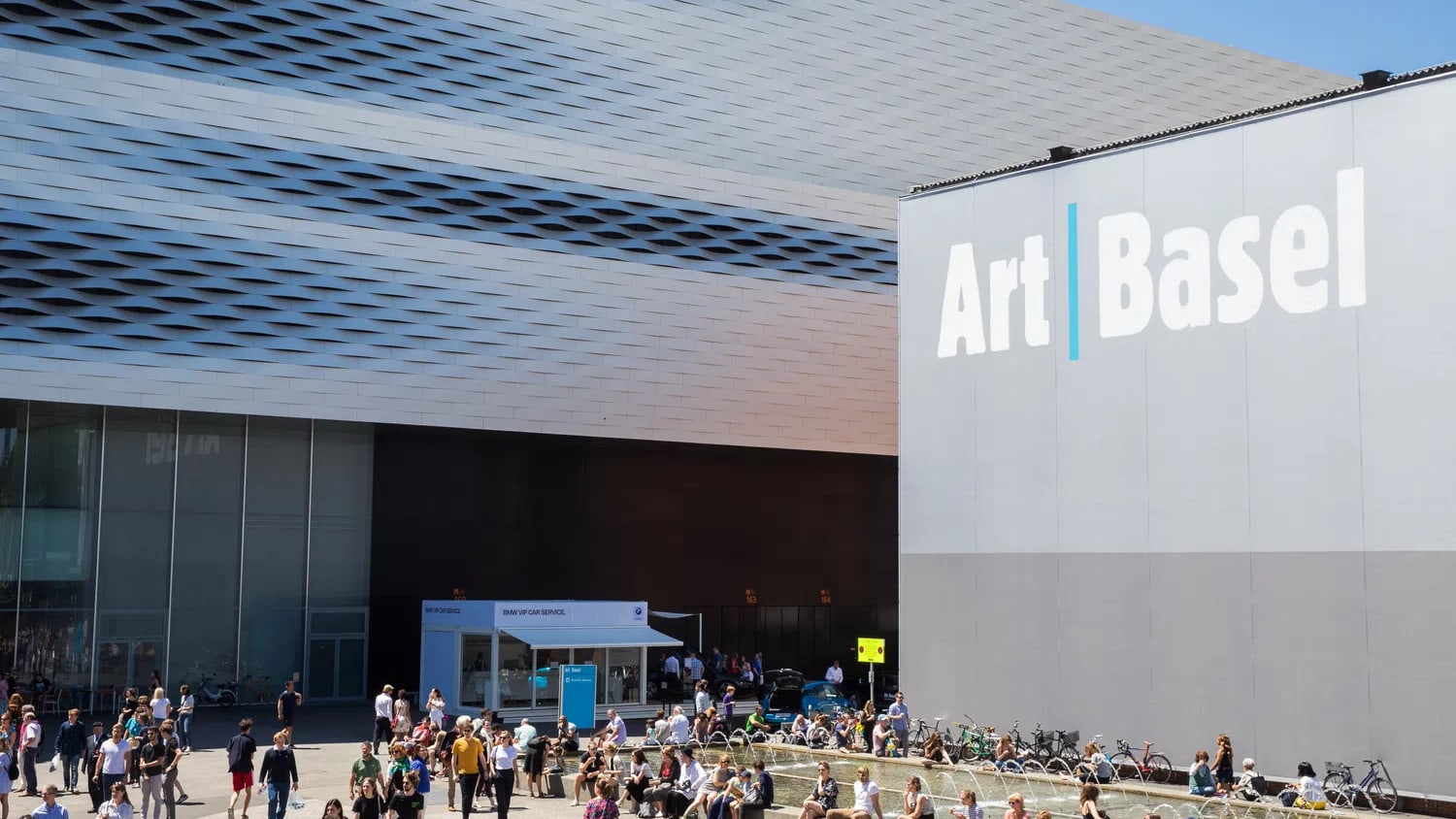 A photograph of the modern Messe Basel exhibition hall building under a clear blue sky, featuring a textured, lattice-like metallic facade on the left and a smooth grey exterior wall on the right prominently displaying the "Art Basel" logo with a blue vertical line separating the words.