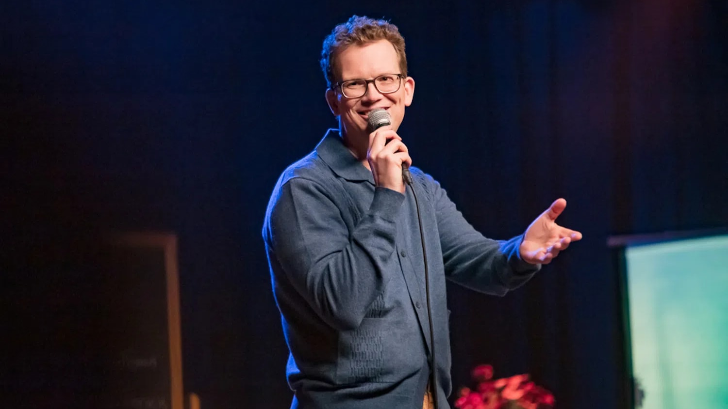 Popular internet educator and author Hank Green stands mid-gesture under vibrant stage lighting during a performance of his debut stand-up comedy special, Pissing Out Cancer, which premiered via Dropout Presents. Wearing his signature thick-rimmed glasses, a structured slate-blue buttoned cardigan polo, and casual tan trousers, he smiles broadly while holding a corded microphone close to his mouth with his right hand and extending his left hand outward to engage with the audience.