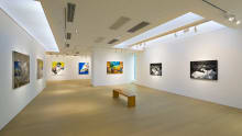 Tang Contemporary Art Gallery exhibition space featuring contemporary paintings on display under bright gallery lighting. An expansive, minimalist art gallery interior features light wood floors and clean white walls illuminated by modern recessed ceiling lights and track lighting systems. The wide-angle perspective showcases a spacious exhibition hall where vibrant contemporary paintings are spaced out across the walls, leading the viewer's eye toward a central wooden bench and an adjacent hallway that extends deeper into the gallery.