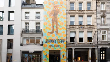 A tall, narrow building facade in an urban streetscape is completely covered by a vibrant, full-height art installation banner by Johnny Depp for his "Halcyon" exhibition. The colorful banner features a light blue background densely patterned with bright orange rose motifs, framing a central, stylized brown silhouette of a humanoid figure with long rabbit-like ears. Below the artwork, the text "JOHNNY DEPP" is printed in large, black, block lettering, directly above the word "HALCYON." A dark rectangular entryway is cut out at the base of the banner, leading into the gallery space. The decorated building is tightly flanked on both sides by traditional, multi-story European city architecture, including a white building with black iron balconies to its left and a classic stone building with rows of windows and white flags to its right, creating a stark contrast between classic street design and contemporary pop-art promotion.