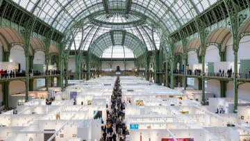 A high-angle, wide-shot photograph captures the vast main exhibition floor of the Art Basel Paris art fair organized inside the sprawling nave of the historic Grand Palais in Paris, France. The massive space is covered by an ornate, vaulted glass and green-painted iron dome roof structure that floods the interior with natural light. Dozens of temporary, white-walled exhibition booths are arranged in a dense, geometric grid across the ground floor, creating corridors filled with visitors browsing contemporary artwork and sculptures. A long, straight central aisle cuts through the middle of the floor layout, packed with people walking between the various gallery displays. Surrounding the main floor, elevated perimeter balconies with green iron railings wrap around the structure, also populated with visitors overlooking the event. Large informational banners hang vertically from the upper railings on either side, while modern artwork, including vibrant abstract paintings and multi-colored wall hangings, is visible inside several open-fronted booths below.