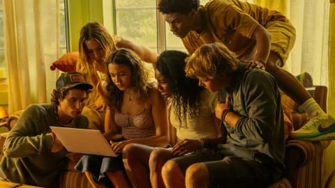A group of six teenagers from the Netflix series Outer Banks are gathered tightly together in a warm, yellow-toned living room, looking intensely at a silver laptop screen. On the far left, a young man wearing a trucker hat sits on the floor leaning inward. Next to him, a young woman with wavy brown hair holds the laptop on her lap. Behind them stands another young woman leaning over the back of the sofa. In the center, a young woman with dark curly hair watches the screen closely, while a young man with shaggy blonde hair sits next to her, leaning his torso forward. Above them, a young man in a yellow shirt and shorts climbs over the back of the sofa, leaning completely over the group to catch a glimpse of the screen.