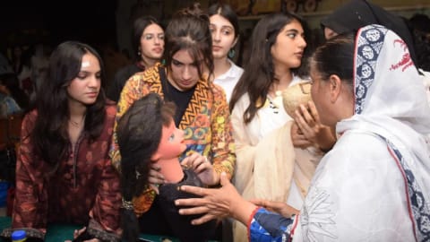 A group of female students at the National College of Arts (NCA) Rawalpindi gathers closely around a table during an interactive puppetry workshop. An elderly female instructor, wearing a traditional white patterned dupatta, demonstrates the mechanics of a dark-haired puppet with a pink face. The students, dressed in diverse casual and traditional South Asian attire, look on with expressions of focused curiosity and engagement.