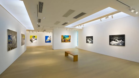 Tang Contemporary Art Gallery exhibition space featuring contemporary paintings on display under bright gallery lighting. An expansive, minimalist art gallery interior features light wood floors and clean white walls illuminated by modern recessed ceiling lights and track lighting systems. The wide-angle perspective showcases a spacious exhibition hall where vibrant contemporary paintings are spaced out across the walls, leading the viewer's eye toward a central wooden bench and an adjacent hallway that extends deeper into the gallery.