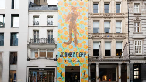 A tall, narrow building facade in an urban streetscape is completely covered by a vibrant, full-height art installation banner by Johnny Depp for his "Halcyon" exhibition. The colorful banner features a light blue background densely patterned with bright orange rose motifs, framing a central, stylized brown silhouette of a humanoid figure with long rabbit-like ears. Below the artwork, the text "JOHNNY DEPP" is printed in large, black, block lettering, directly above the word "HALCYON." A dark rectangular entryway is cut out at the base of the banner, leading into the gallery space. The decorated building is tightly flanked on both sides by traditional, multi-story European city architecture, including a white building with black iron balconies to its left and a classic stone building with rows of windows and white flags to its right, creating a stark contrast between classic street design and contemporary pop-art promotion.